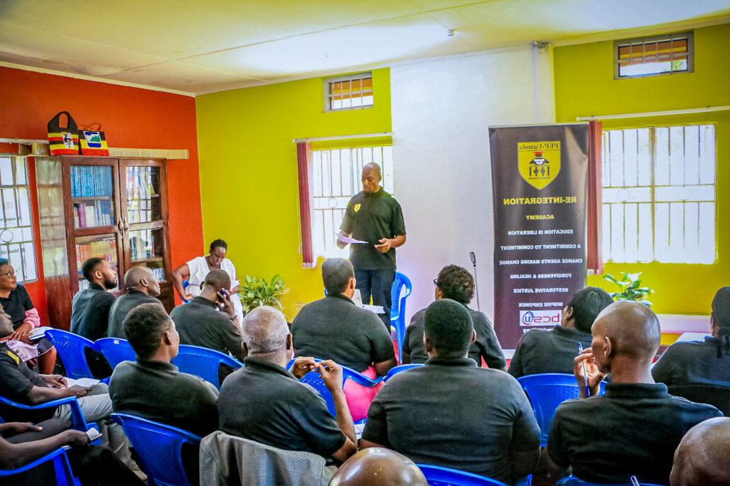 Kenneth Butler stands in front of a class while reading from a paper in his hand. Butler wears a black polo shirt with the yellow logo for PEP-Uganda on the upper left corner. To his right is a black banner for the Reintegration Academy with the statement “Education is liberation, 对承诺的承诺, 变革推动者做出变革, 宽恕 & healing, restorative justice, inspire empower.” 