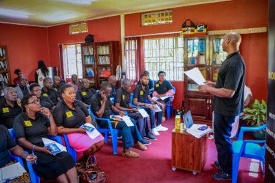 A classroom full of people in black polo shirts with the yellow logo for PEP-Uganda sit at attention as Butler presents to them. 